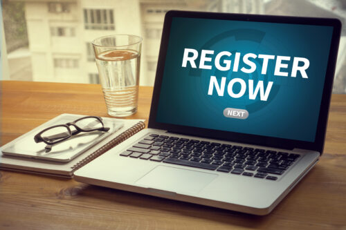 Ein Laptop auf einem Tisch zeigt den Schriftzug „Register Now“ auf dem Bildschirm.